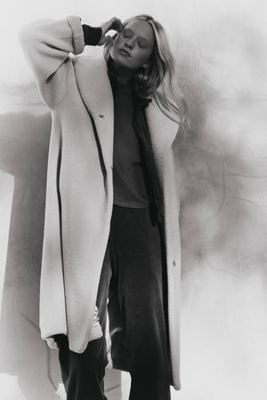 Black and white photo of a person standing against a textured wall, wearing the ELEANOR SHERPA REVERSIBLE COAT by Velvet by Graham & Spencer and loose pants, raising one hand to their head.