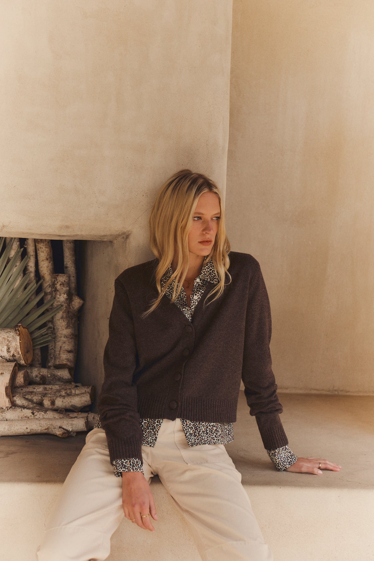 A blonde woman sits on a ledge near a fireplace in a neutral-toned room, wearing the ETNA CARDIGAN by Velvet by Graham & Spencer over a patterned blouse and light pants.-39814881542337
