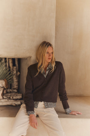 A blonde woman sits on a ledge near a fireplace in a neutral-toned room, wearing the ETNA CARDIGAN by Velvet by Graham & Spencer over a patterned blouse and light pants.