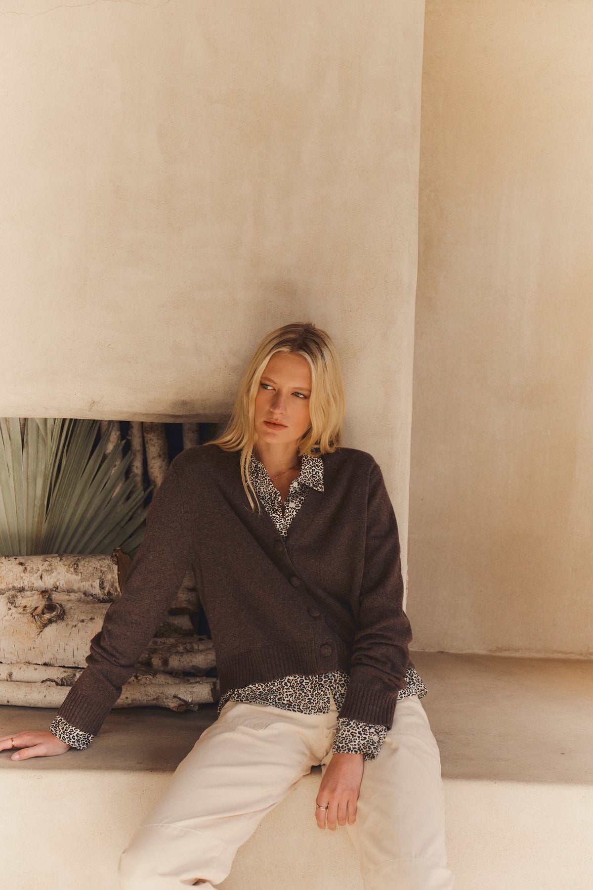 A woman with blonde hair sits against a light wall, wearing the ETNA CARDIGAN by Velvet by Graham & Spencer over a patterned shirt and beige pants. Dried palm leaves and logs behind her complete the cozy look.-39814881476801