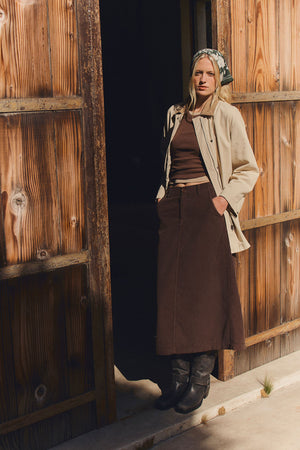A woman stands in a wooden doorway with her hands in her pockets, wearing a beige jacket, brown top, black boots, patterned headscarf, and the FIA CORDUROY SKIRT by Velvet by Graham & Spencer.