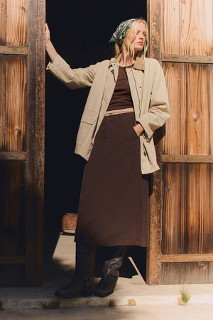 A person stands in a wooden doorway, wearing Velvet by Graham & Spencer's ROSA JACKET—an oversized beige piece styled with a green headscarf, brown top and skirt, and black boots, creating an effortlessly chic everyday look.