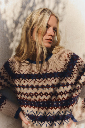 A woman with long blonde hair wears the MAKENA ALPACA SWEATER by Velvet by Graham & Spencer, standing against a light wall with dappled shadows.