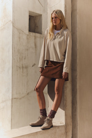 A woman with long blonde hair wears a beige long-sleeve shirt, maroon socks, beige ankle boots, and the LIZA CORDUROY SKIRT by Velvet by Graham & Spencer, standing against an off-white concrete wall.