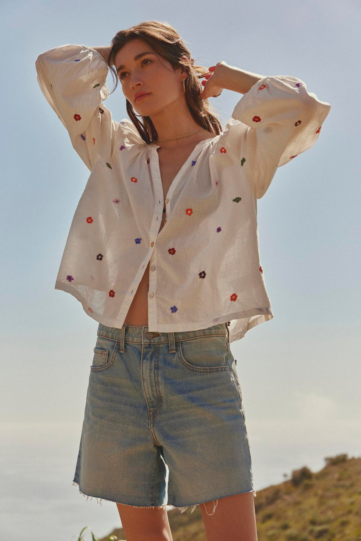 Woman in a Velvet by Graham & Spencer ARETHA EMBROIDERED BOHO TOP with floral embroidery and denim shorts standing outdoors with hands on her head.-36532838924481