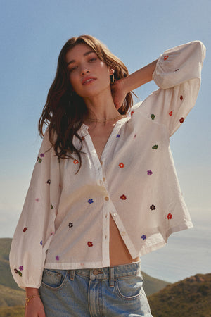 A woman stands outdoors, wearing the ARETHA EMBROIDERED BOHO TOP by Velvet by Graham & Spencer, paired with blue jeans. She has long brown hair and one arm raised, touching the back of her head.