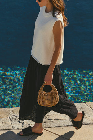 A person in a sleeveless white top, long black skirt, and black sandals walks by a pool, carrying the MICRO SLOUCH BAG by Velvet by Graham & Spencer—a chic paper straw accessory perfect for their summery look.