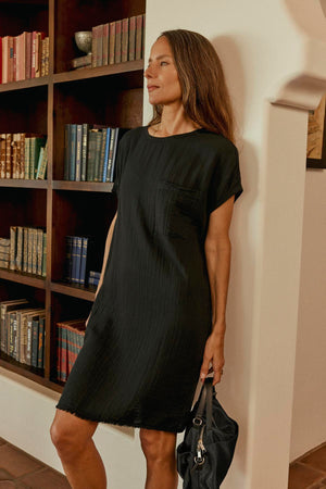 A woman in a black dress stands by a bookshelf, elegantly holding the NYLON CROSSBODY BAG from Velvet by Graham & Spencer.