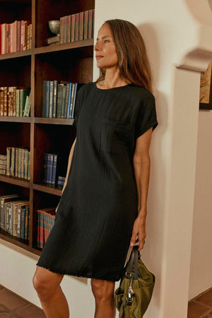 A woman in a HANNA DRESS by Velvet by Graham & Spencer with a cocoon silhouette stands by a bookshelf, holding a green handbag.