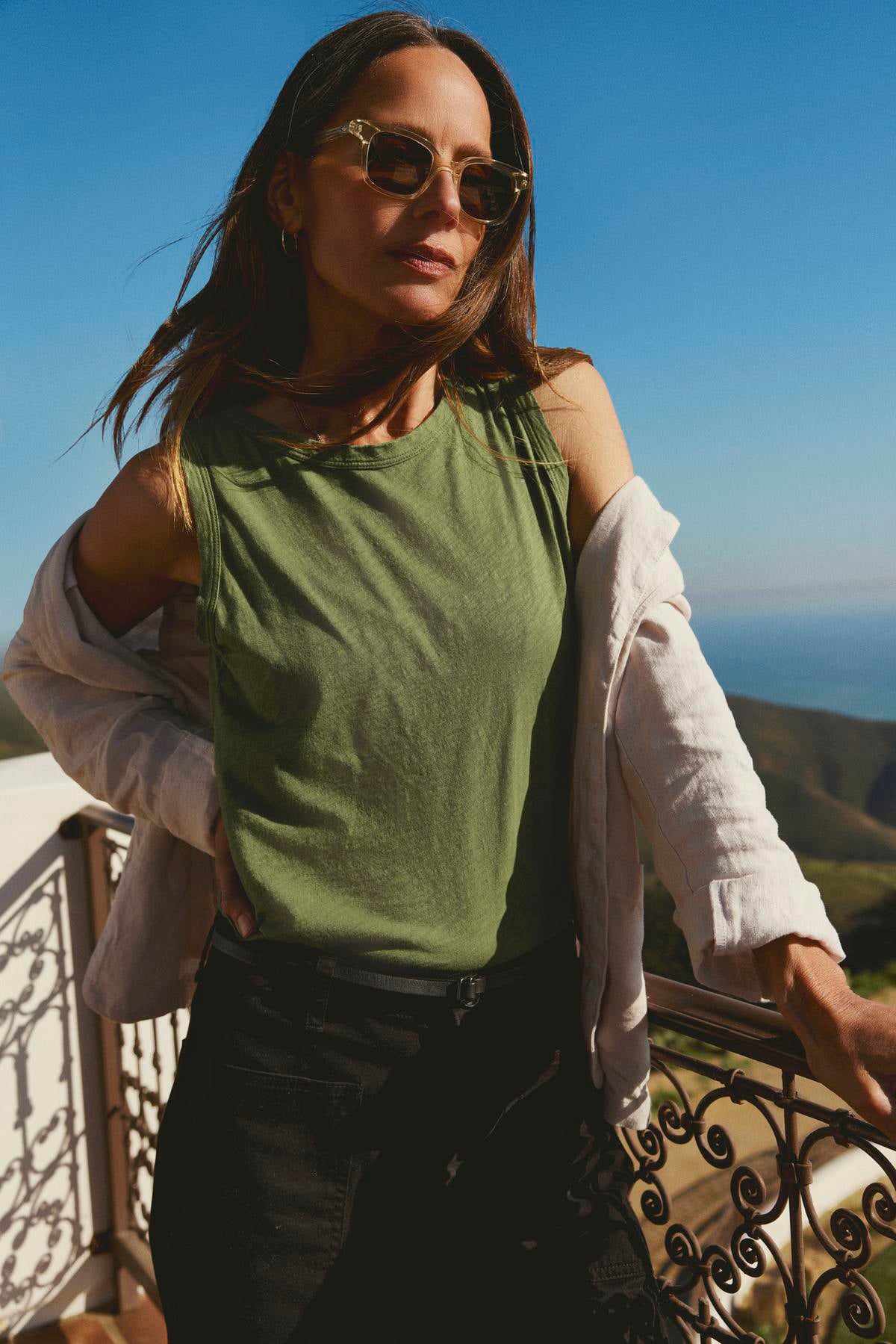   A woman in sunglasses, a Velvet by Graham & Spencer TAURUS COTTON SLUB TANK, and white shirt stands by a railing with a coastal view in the background. 