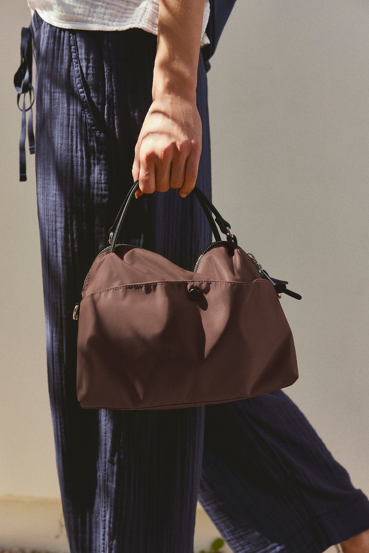   A person is holding the NYLON CROSSBODY BAG by Velvet by Graham & Spencer, featuring a brown design with black handles for everyday use, while wearing navy blue pants. 