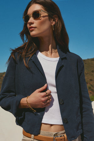 A woman wearing sunglasses, a Navy Notched Collar Blazer by Velvet by Graham & Spencer, and a white top stands outdoors with a clear blue sky in the background.