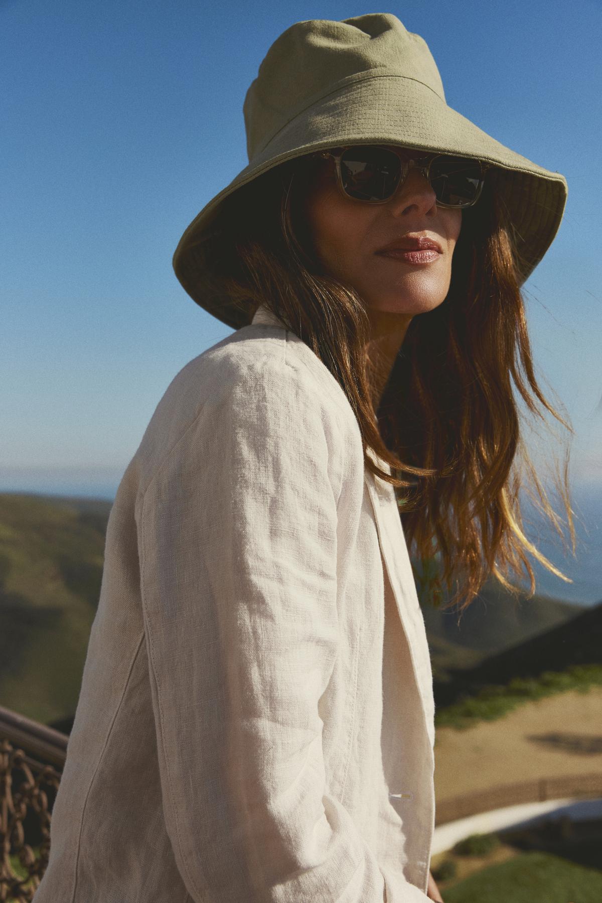   Woman in a Hazel Bucket Hat by Freya and sunglasses looking over her shoulder, with a scenic hilly background under a clear sky. 