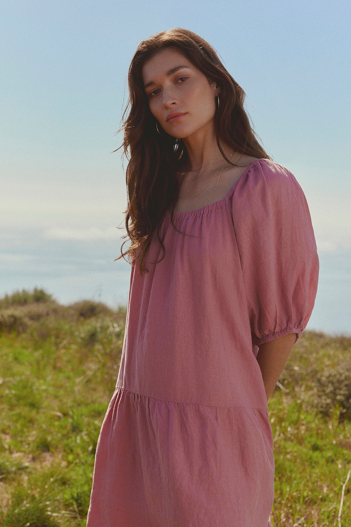 A woman in a pink Velvet by Graham & Spencer IRINA LINEN TIERED DRESS standing in a field with the ocean in the background, gazing towards the camera.-36783641395393