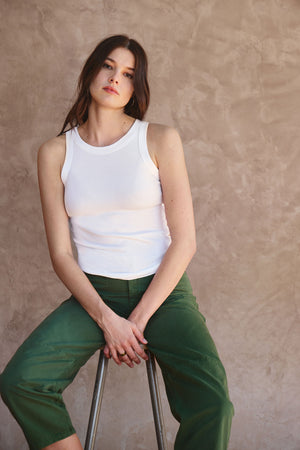 A woman with long brown hair wears the CRUZ TANK TOP by Velvet by Jenny Graham and green pants, sitting on a stool against a beige textured wall—an effortless capsule wardrobe look.