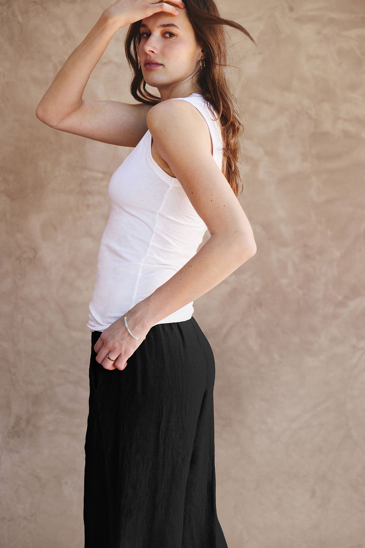   A woman stands sideways against a beige textured wall, wearing a white sleeveless top and Velvet by Graham & Spencer's LOLA LINEN PANT—black, pull-on trousers with a relaxed leg—one hand raised to her forehead. 
