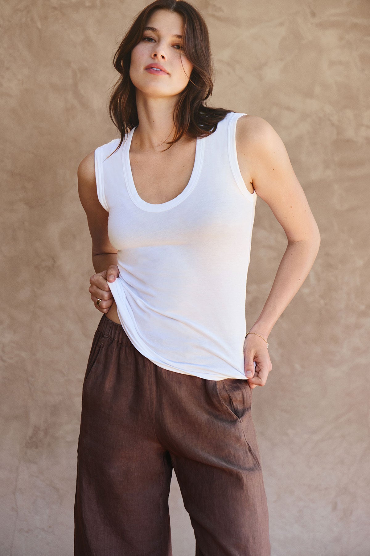   A woman wearing Velvet by Graham & Spencer's LOLA LINEN PANT, a brown lightweight linen pull-on trouser with an elastic waist, stands before a beige textured background, holding the hem of her white sleeveless top. 