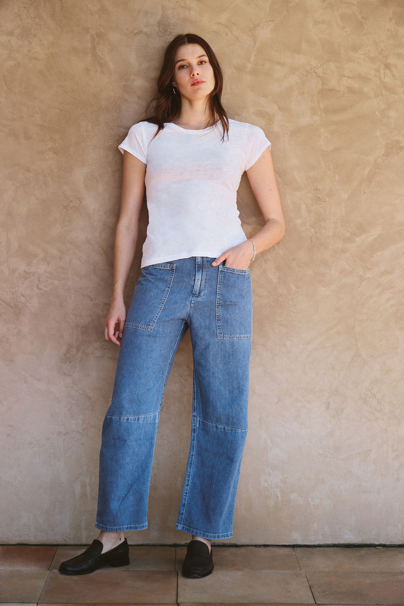 A person stands against a textured beige wall, wearing the ODELIA TEE by Velvet by Graham & Spencer—a white premium cotton t-shirt—paired with blue wide-leg jeans and black slip-on shoes.