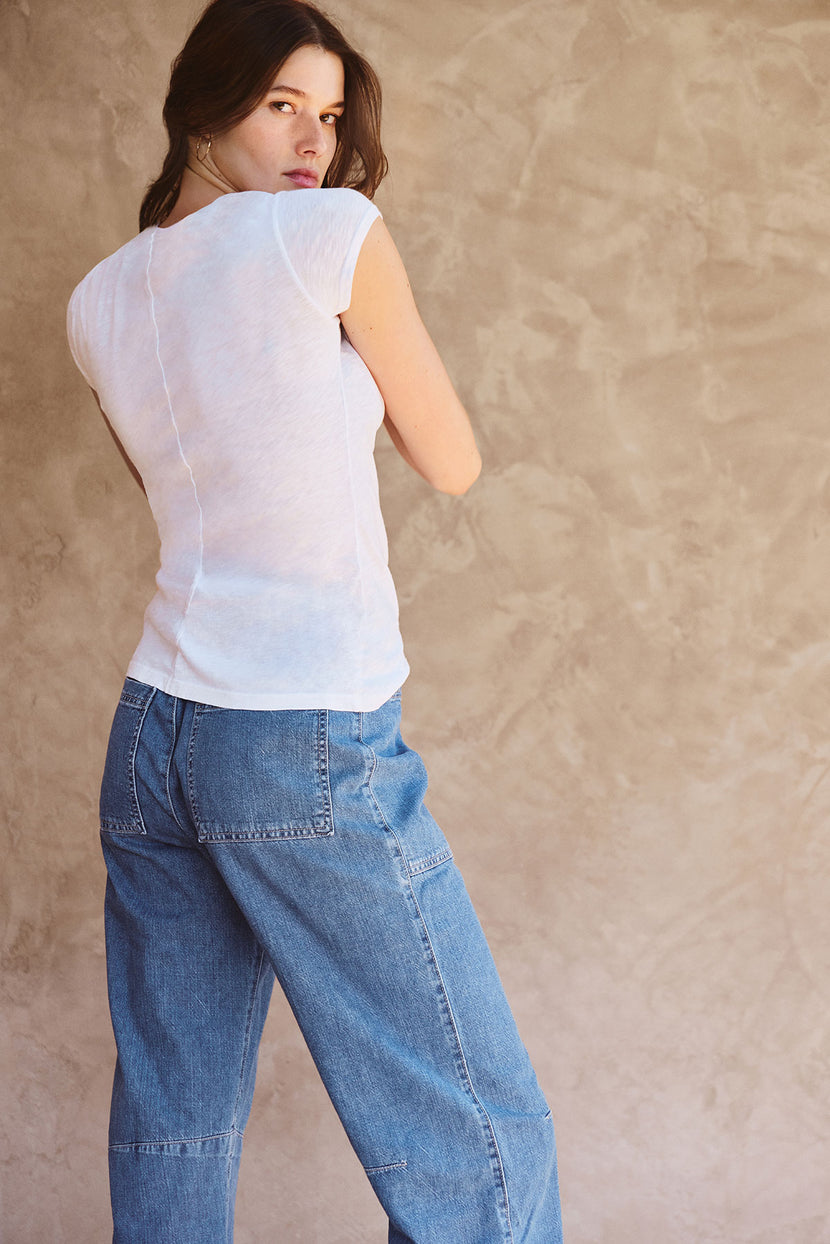 A woman with long brown hair wears the ODELIA TEE by Velvet by Graham & Spencer, a white short-sleeve crew neck, paired with blue jeans, and stands with her back to a beige textured wall.