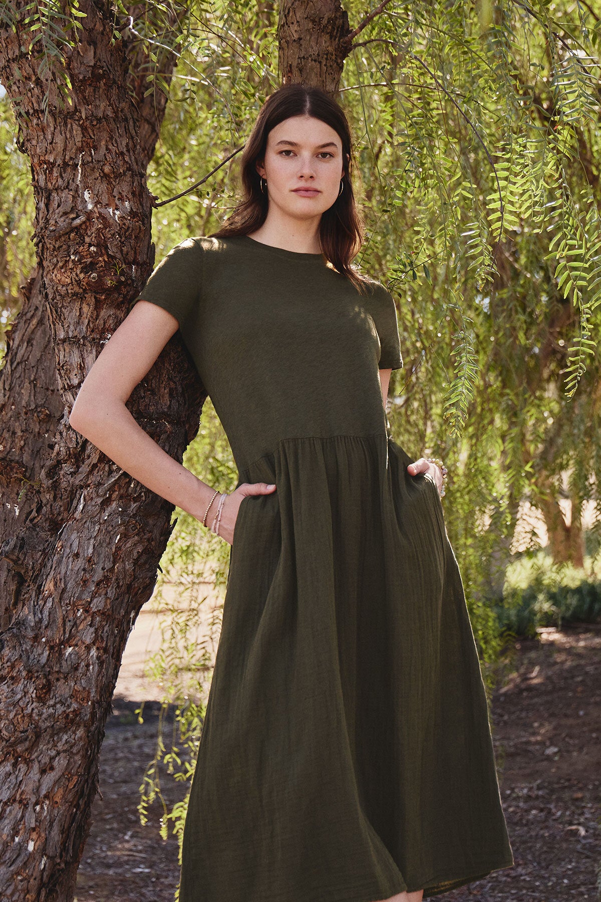   A woman in the ITZA DRESS by Velvet by Graham & Spencer stands by a tree with hands in pockets, surrounded by foliage. The soft fabric of her green midi cotton slub dress complements the natural scenery. 