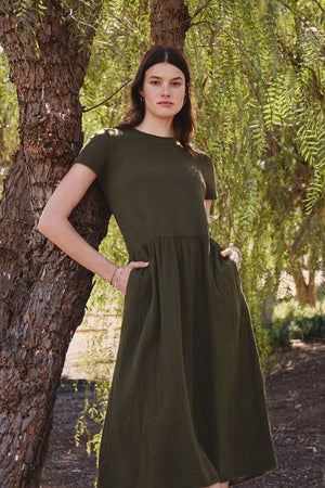 A woman in the ITZA DRESS by Velvet by Graham & Spencer stands by a tree with hands in pockets, surrounded by foliage. The soft fabric of her green midi cotton slub dress complements the natural scenery.