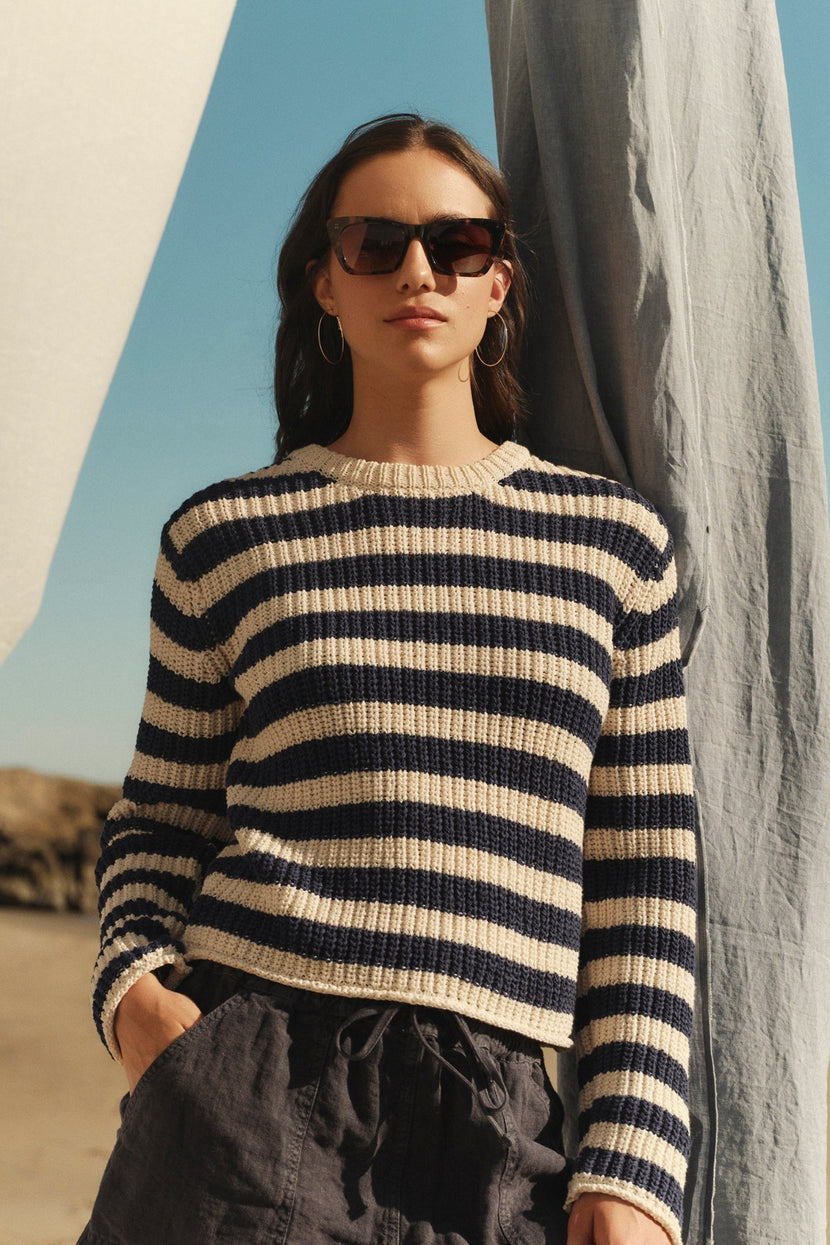 A woman wearing a MAXINE SWEATER from Velvet by Graham & Spencer and sunglasses stands confidently in front of a draped fabric background under a clear sky.