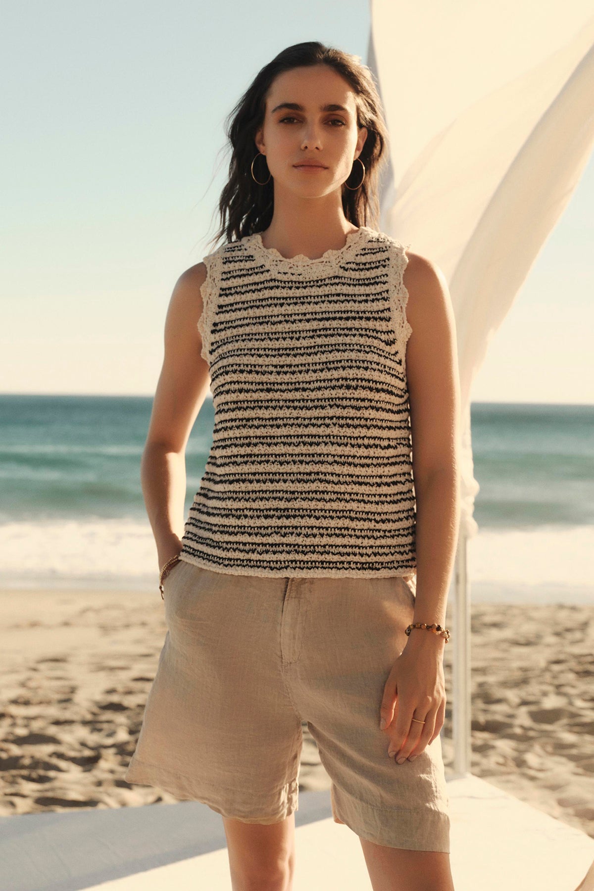 Woman in a sleeveless Sophie sweater vest by Velvet by Graham & Spencer and shorts standing on a sandy beach with the ocean in the background and a white fabric element on the left.-36910013776065