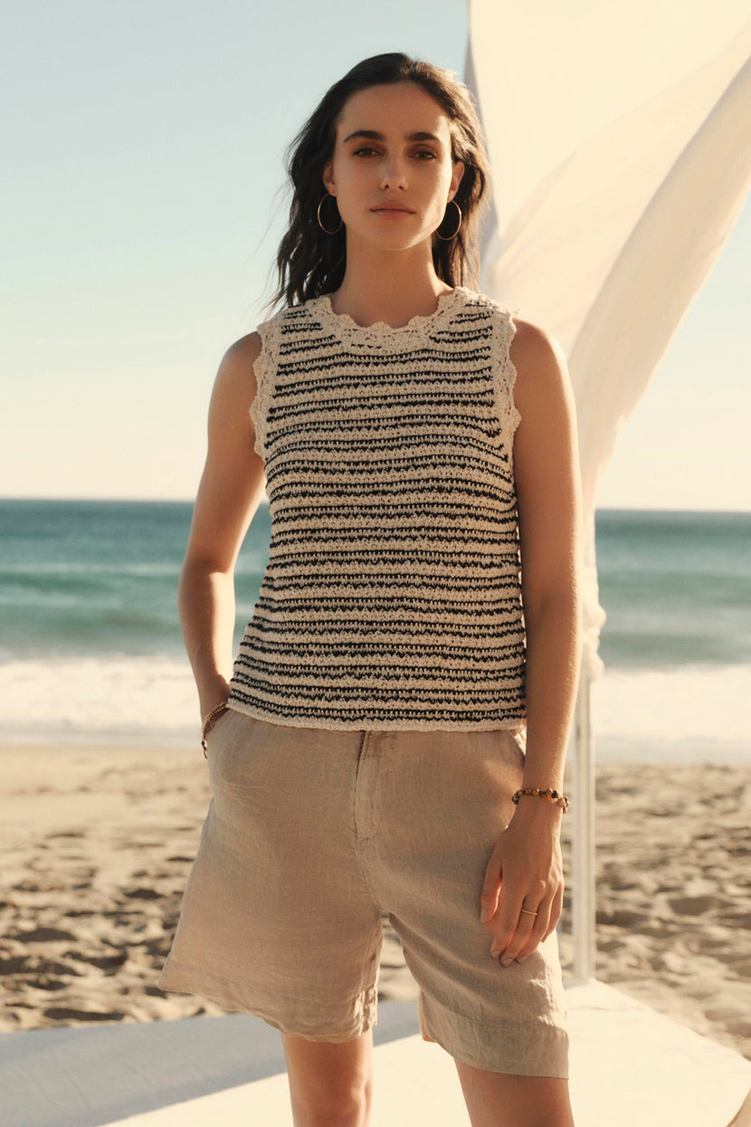 Woman in a sleeveless Sophie sweater vest by Velvet by Graham & Spencer and shorts standing on a sandy beach with the ocean in the background and a white fabric element on the left.