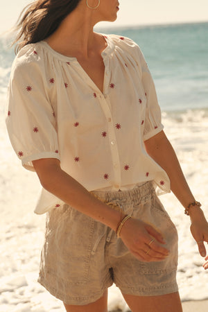 Woman in a white blouse with red floral embroidery, featuring a split neck, and beige shorts, walking on a sunny beach wearing the AMIRA TOP by Velvet by Graham & Spencer.