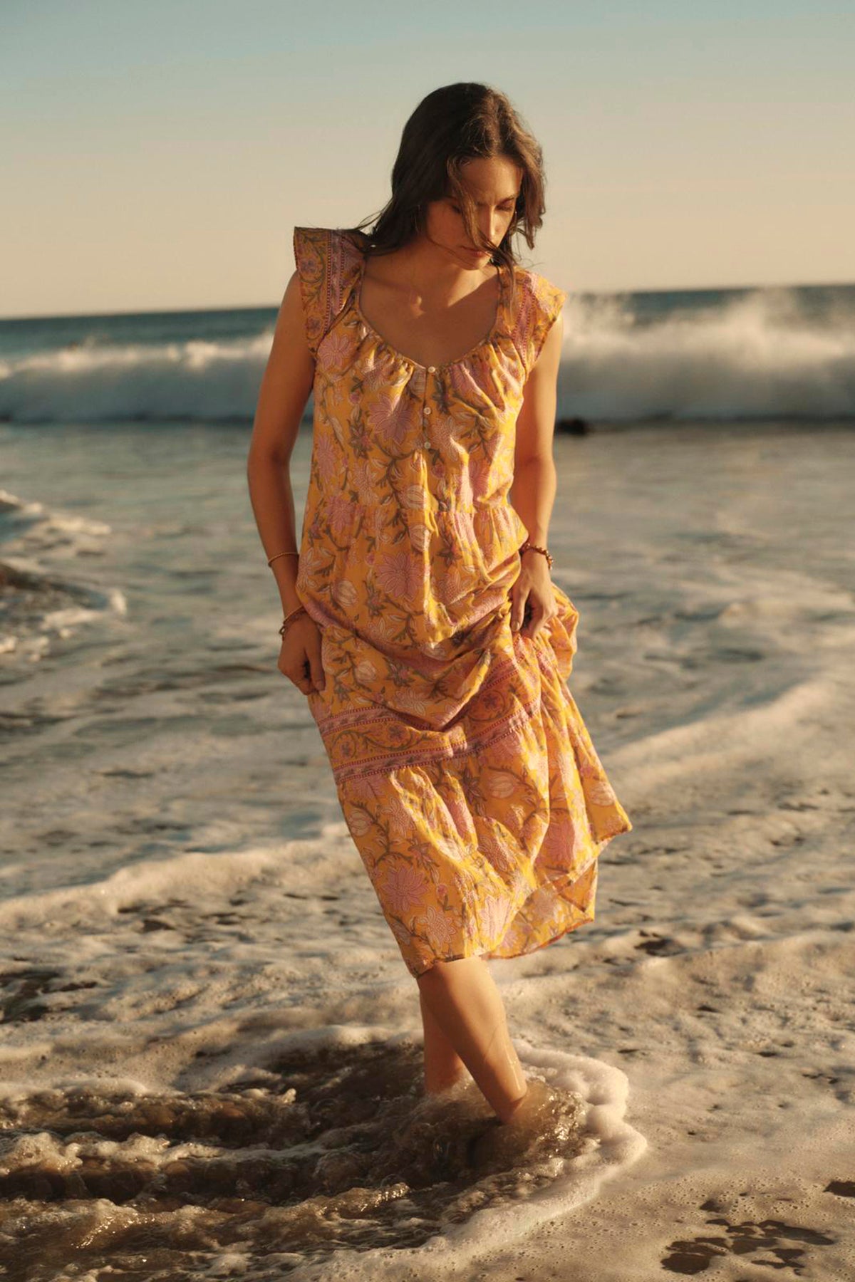   A woman wades through shallow ocean water at the beach, with waves breaking in the background, wearing the JAZLYN DRESS by Velvet by Graham & Spencer. This pink and yellow printed cotton voile midi-length dress is perfect for your summer wardrobe, effortlessly combining style and comfort. 