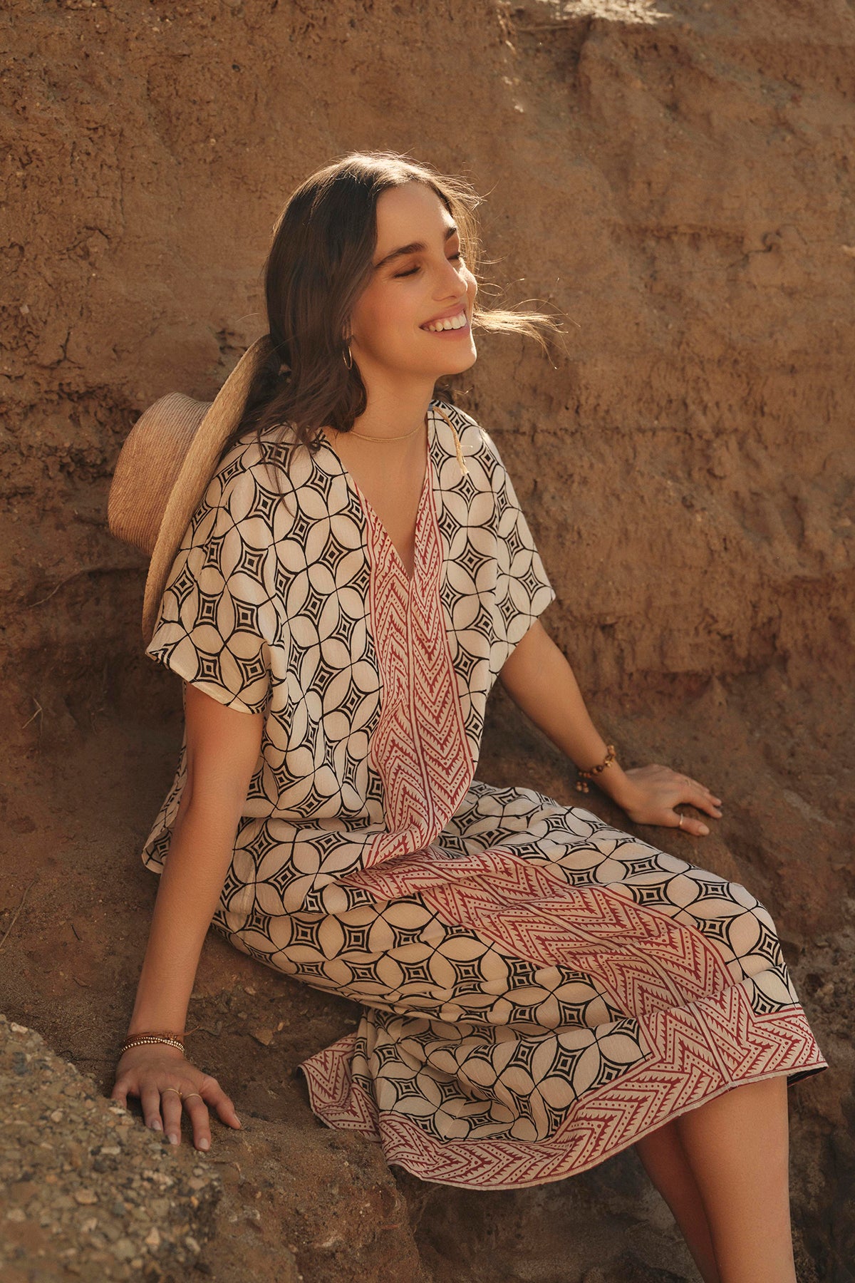 A woman in a lightweight cotton Velvet by Graham & Spencer Odessa kaftan dress smiles while sitting on a rocky terrain, enjoying the sunlight.-36910025769153