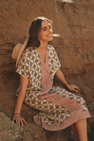 A woman in a lightweight cotton Velvet by Graham & Spencer Odessa kaftan dress smiles while sitting on a rocky terrain, enjoying the sunlight.
