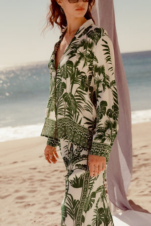 A person wearing the AYLA BUTTON-UP SHIRT by Velvet by Graham & Spencer, featuring a green and white palm print, stands on a sandy beach with the ocean in the background—a true summer essential.