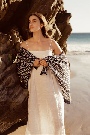 A woman in a Velvet by Graham & Spencer STEPHIE LINEN DRESS and draped patterned shawl stands by a rocky beach, gazing thoughtfully away from the camera.