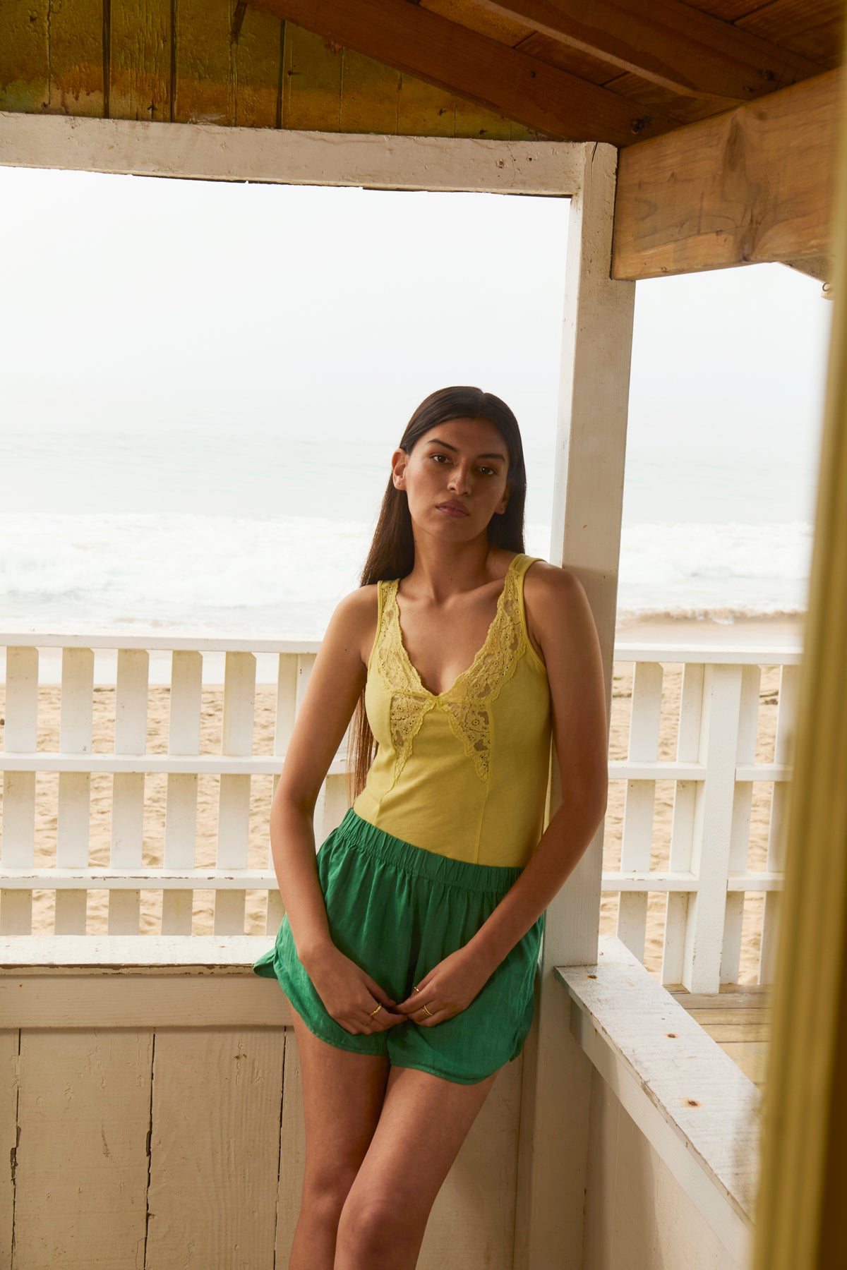 A woman with long hair stands on a beachside porch wearing the yellow HANNAH LACE TANK TOP by Velvet by Graham & Spencer and green shorts, with the ocean and sandy beach in the background.-39119886024897