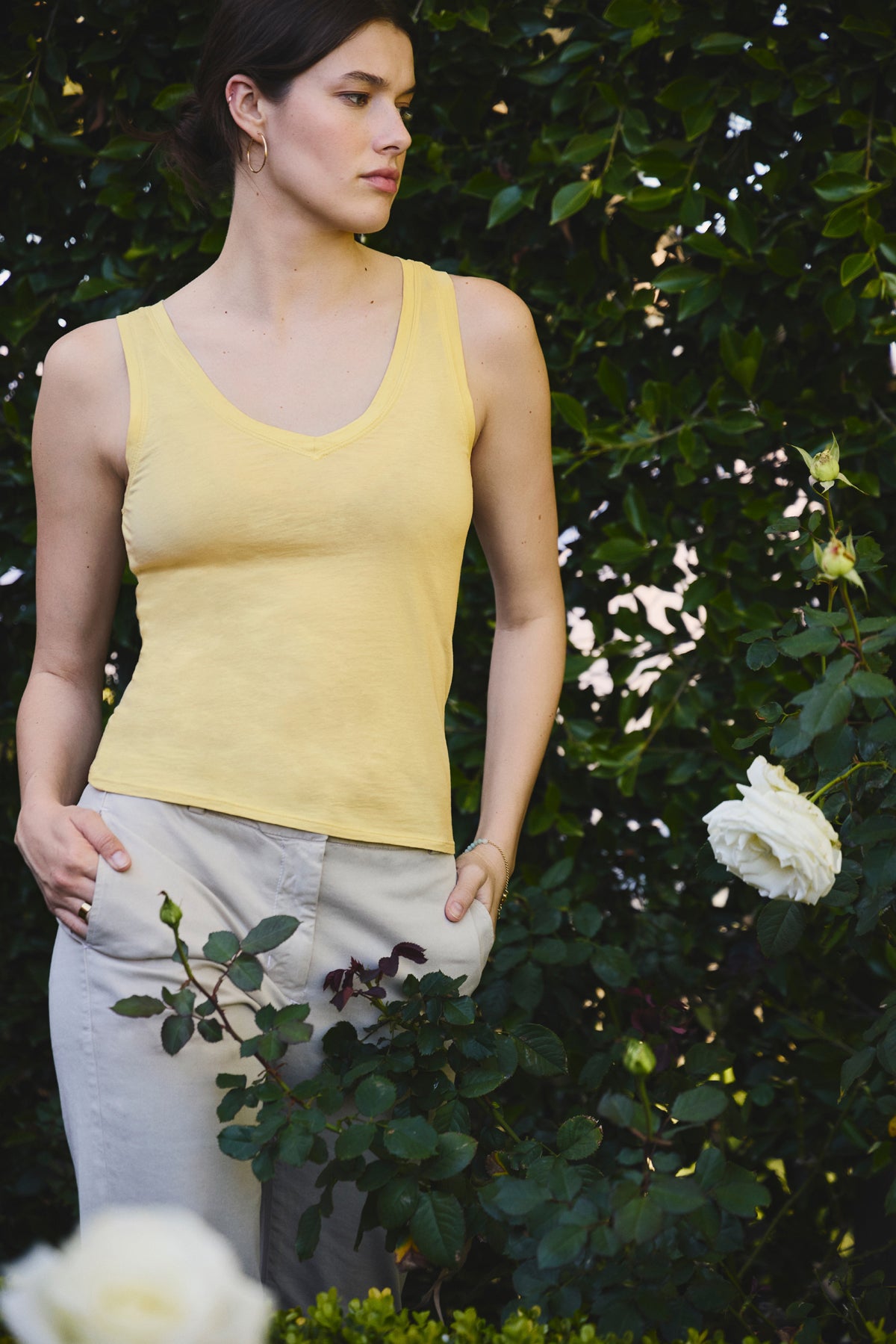 A woman wearing the EVELYN TANK TOP by Velvet by Graham & Spencer and light pants stands outdoors beside a rose bush with white roses, green leaves forming the background.-39096621826241