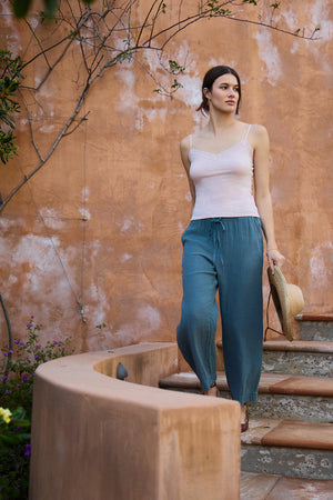 A woman wears the Velvet by Graham & Spencer INDIE LACE CAMI in light pink with loose blue pants, strolling barefoot down stone steps and holding a straw hat, framed by an orange textured wall and climbing plants.