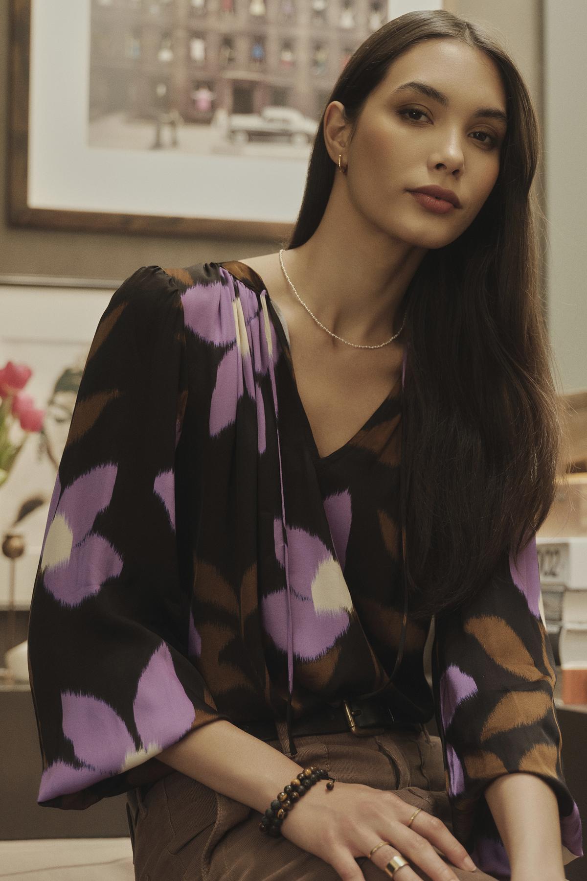 A woman is sitting on a chair in a room, wearing an ISRA PRINTED V-NECK BLOUSE by Velvet by Graham & Spencer with a timeless look and V-neckline.-35416378147009