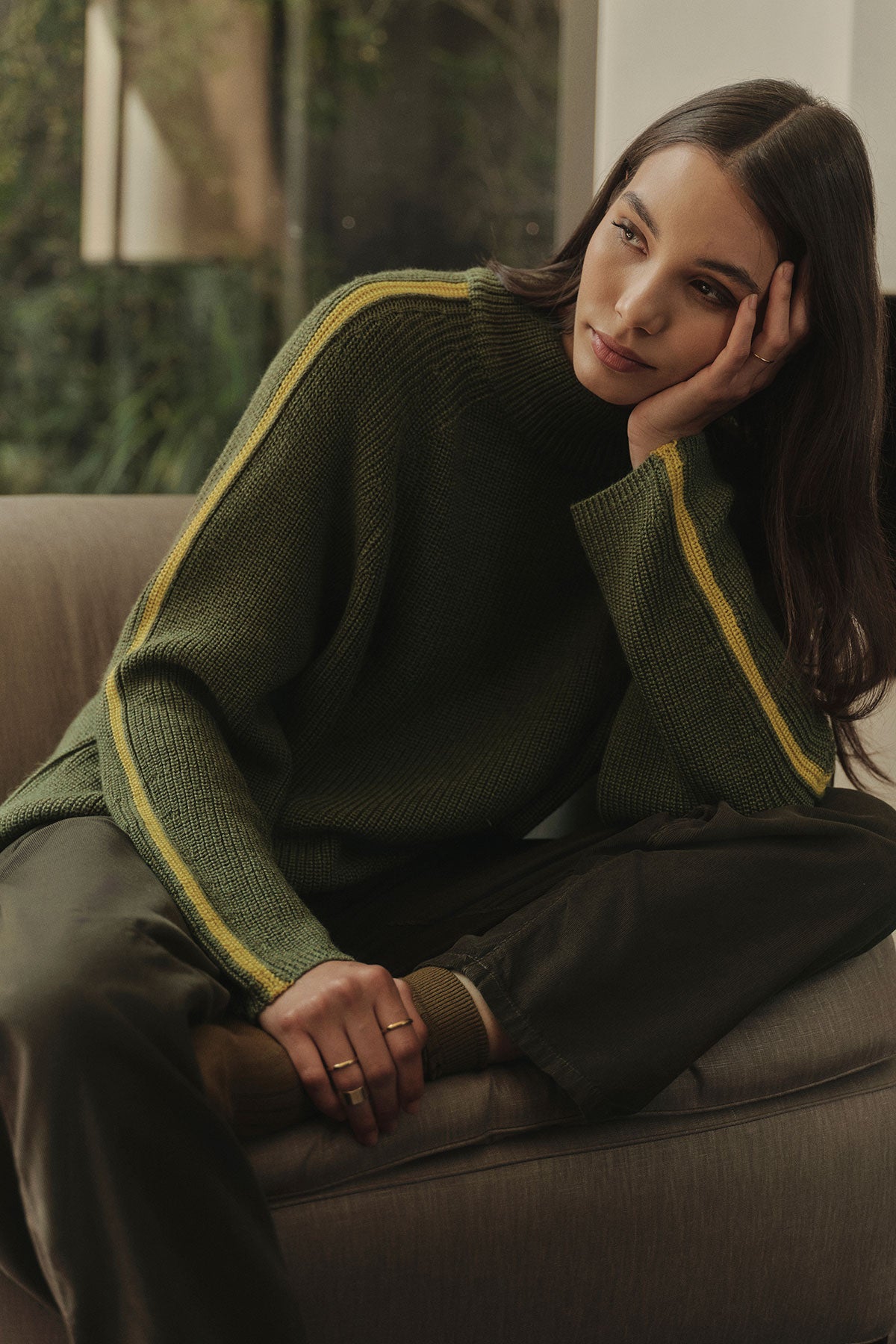   A woman is sitting on a cozy couch in a green Velvet by Graham & Spencer TEAGAN MOCK NECK SWEATER. 