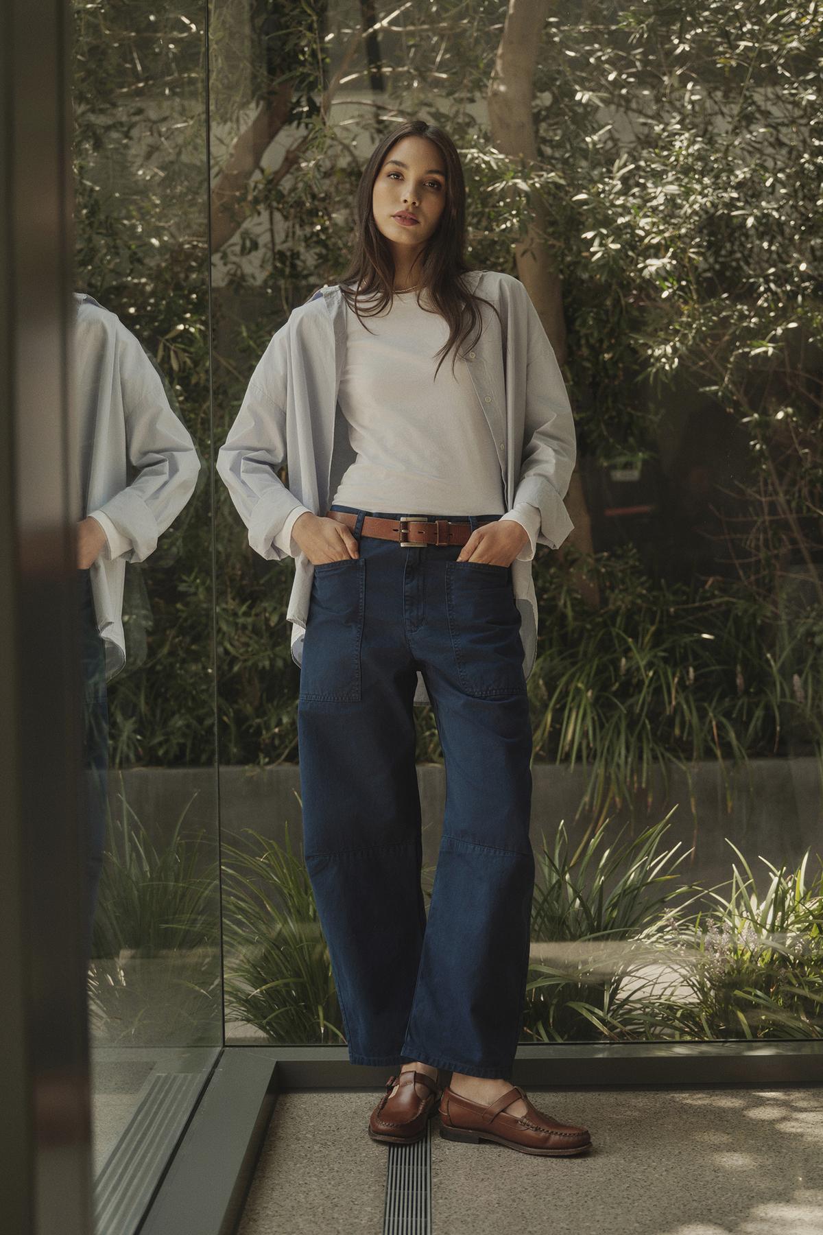   A woman is standing in front of a window wearing Velvet by Graham & Spencer BRYLIE SANDED TWILL UTILITY PANT and a white shirt. 