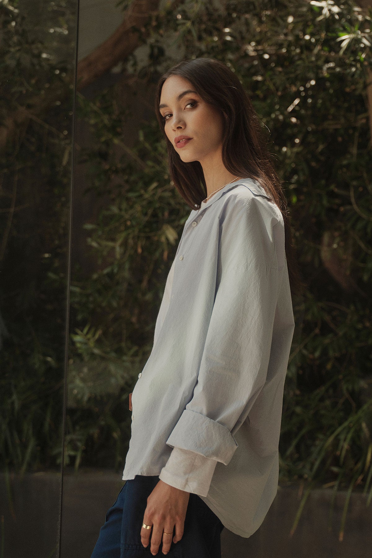   A woman wearing the Velvet by Graham & Spencer Dakota Button-Up Shirt, a classic light blue shirt made of cotton poplin fabric, paired with blue jeans in a modern design. 