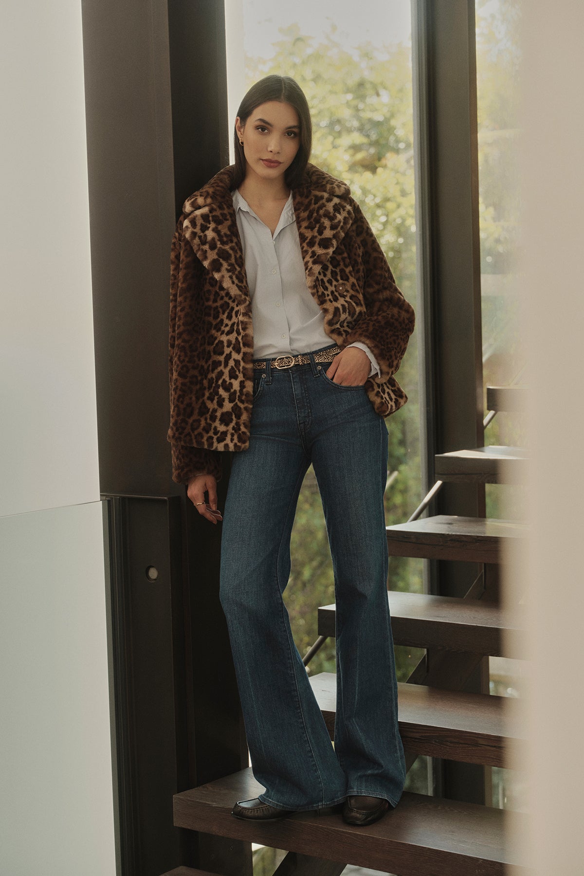A woman standing on a staircase, wearing the AMANI LEOPARD LUX FAUX FUR JACKET by Velvet by Graham & Spencer, a white blouse, and flared jeans, with sunlight filtering through large windows in the background. The ensemble is a perfect addition to any fall wardrobe.-35766170288321