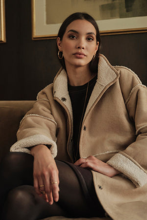 A woman in a relaxed silhouette Albany Lux Sherpa Reversible Jacket sitting on a couch.