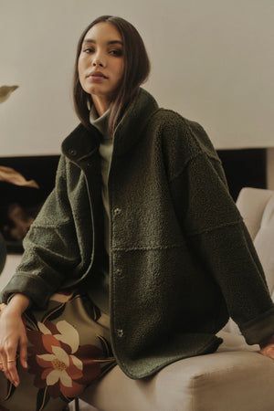 A woman is sitting on a couch in a Velvet by Graham & Spencer ALBANY LUX SHERPA REVERSIBLE JACKET.