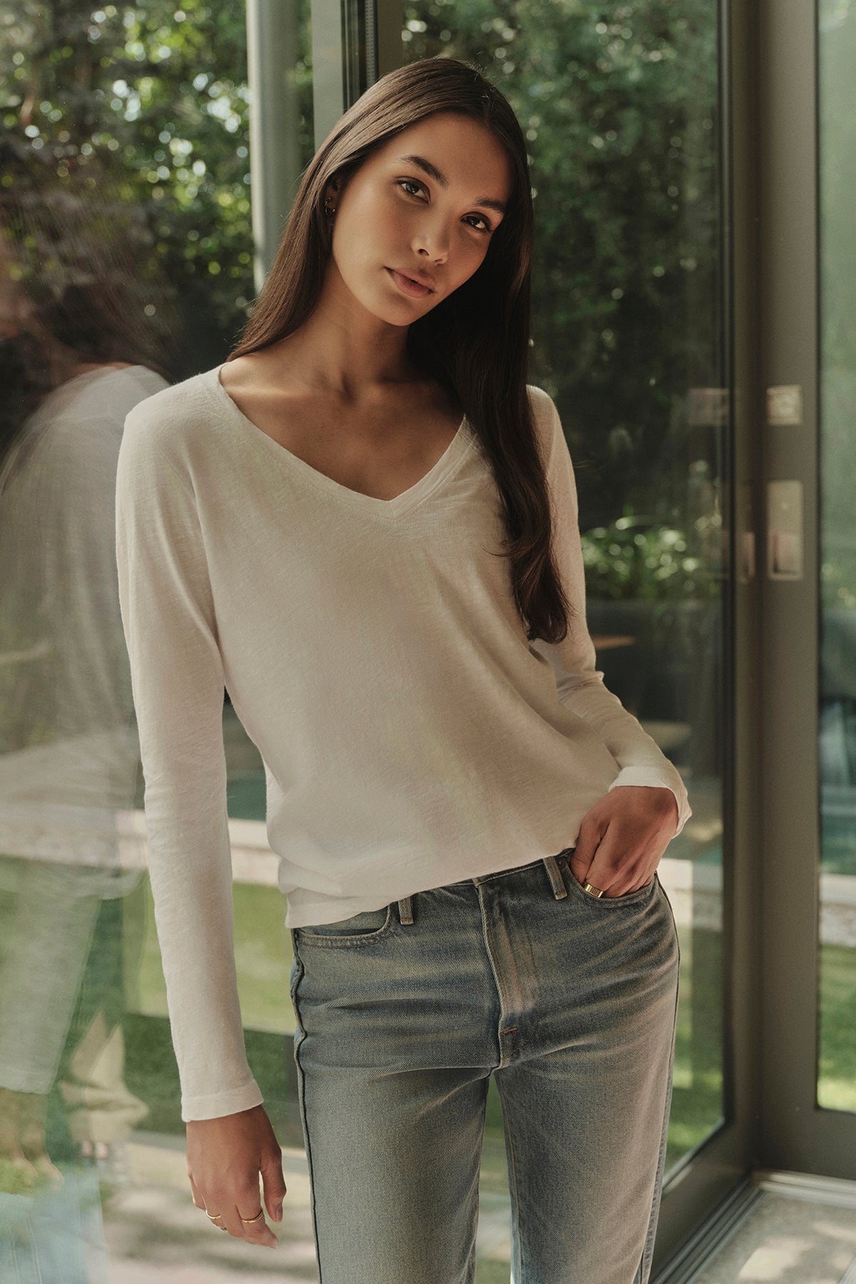   A woman with long brown hair, wearing a white BLAIRE TEE by Velvet by Graham & Spencer and blue jeans, stands in front of a glass door with greenery outside. 