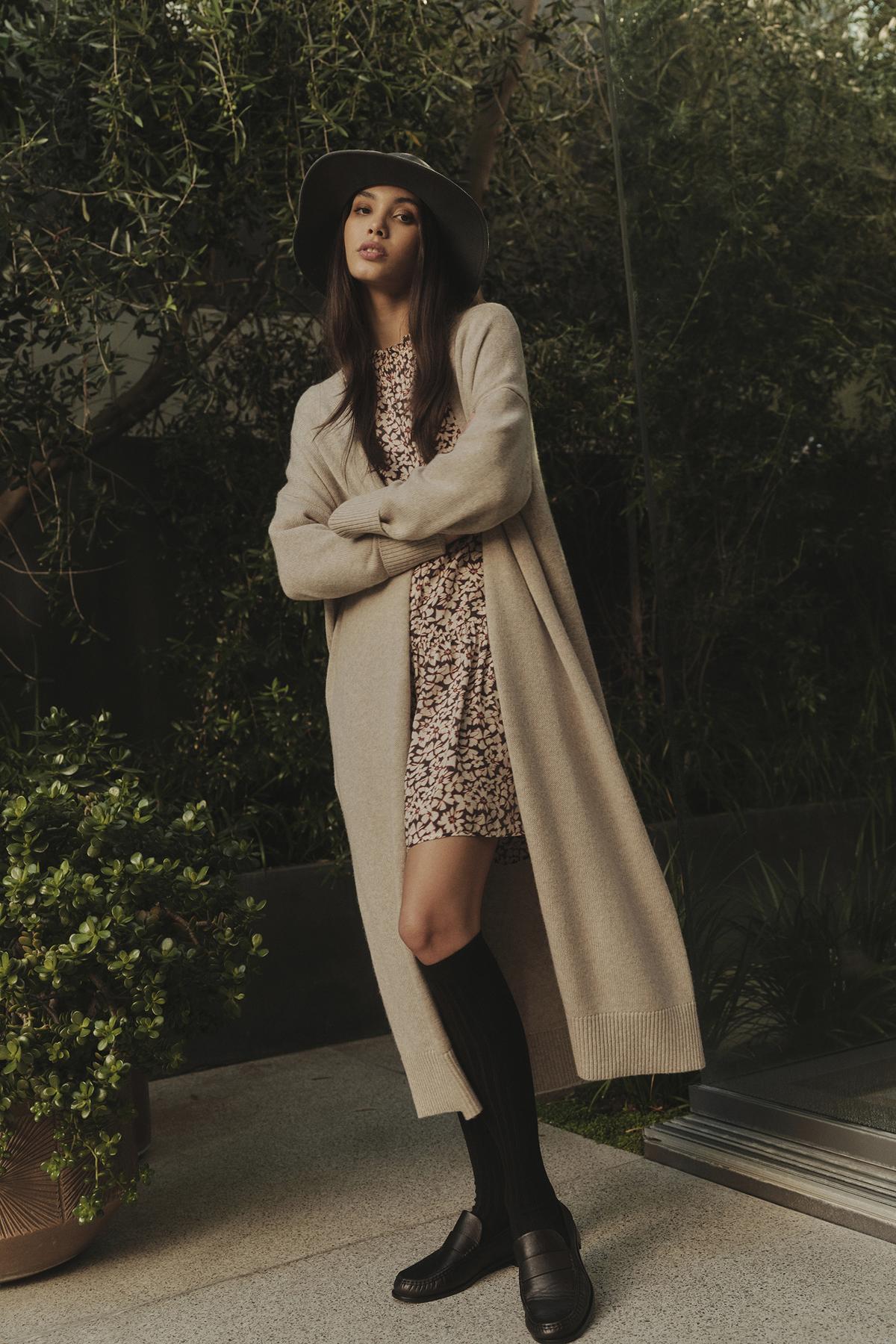 A woman in a beige coat and hat is posing for a photo, showcasing the elegant neckline of her Velvet by Graham & Spencer GENE PRINTED TIERED DRESS.-35445549727937