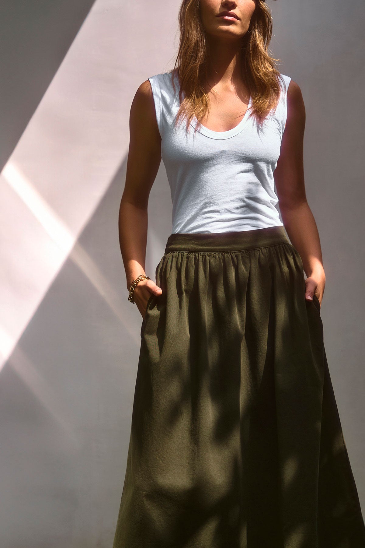   A woman stands under soft, natural light with her hands in her pockets, wearing the ESTINA TANK TOP by Velvet by Graham & Spencer and an olive green skirt for a relaxed, whisper-soft look. 