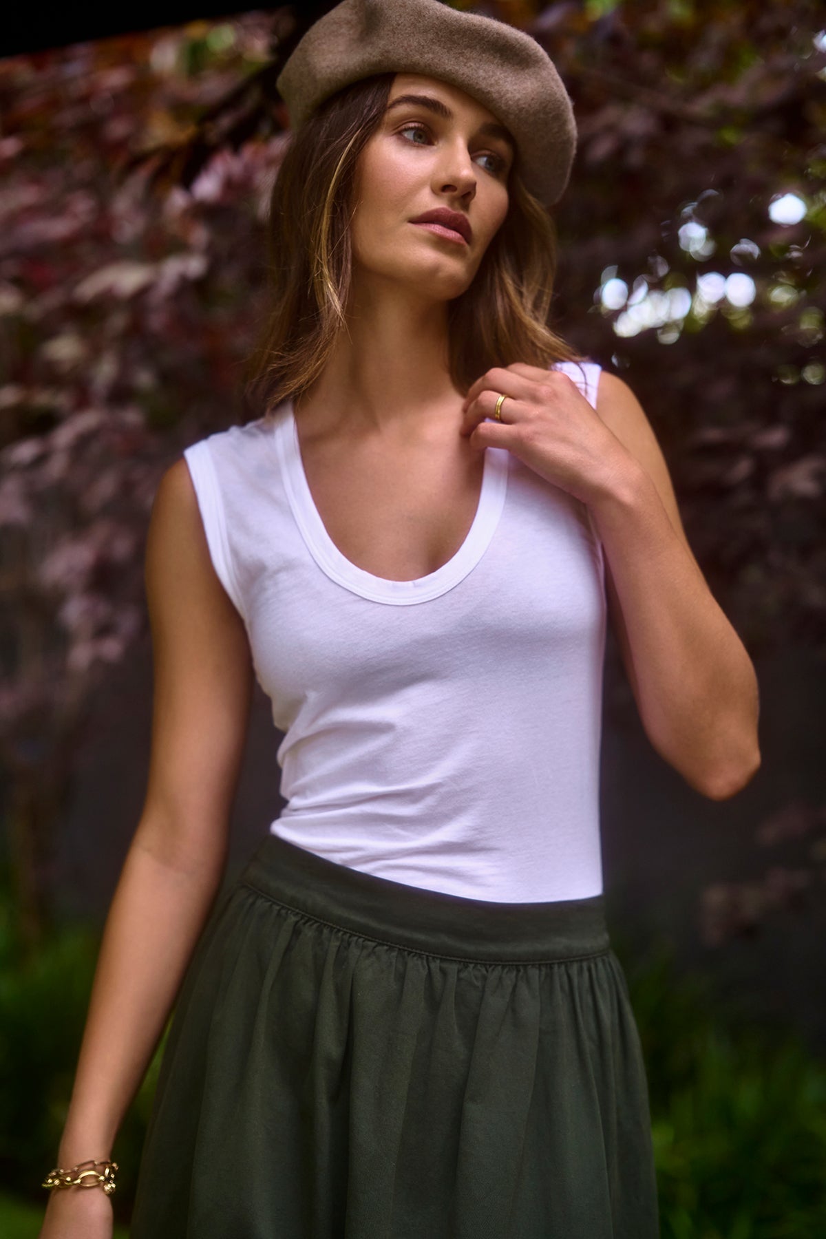   A woman wearing the ESTINA TANK TOP by Velvet by Graham & Spencer in whisper white, paired with a gray beret and a dark green skirt, stands outdoors, touching her hair and looking to the side. 