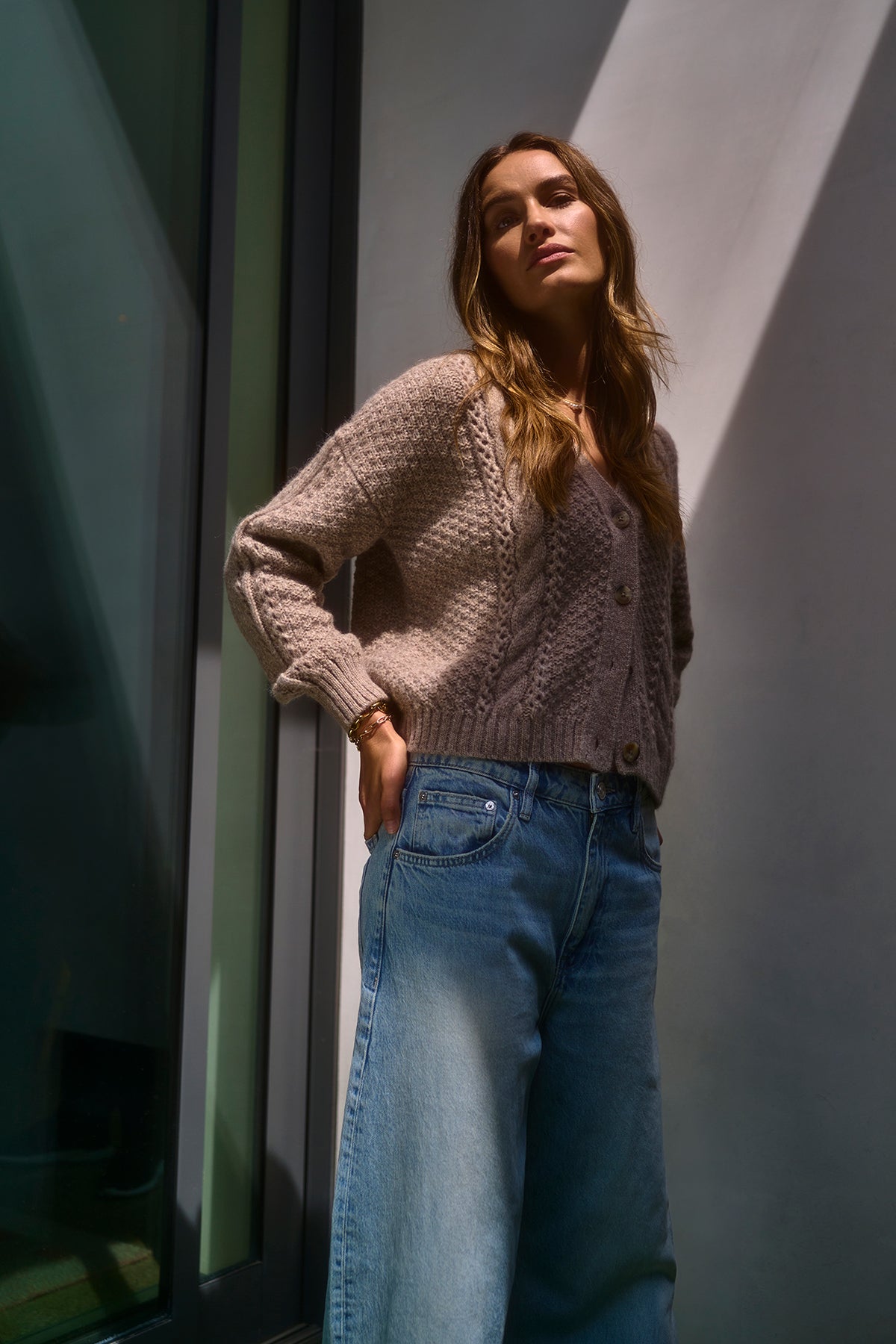   A woman wearing Velvet by Graham & Spencer's IZZY CARDIGAN, a plush alpaca blend with a cable knit pattern, paired with wide-legged jeans stands near a window as light and shadows create a dramatic effect. 