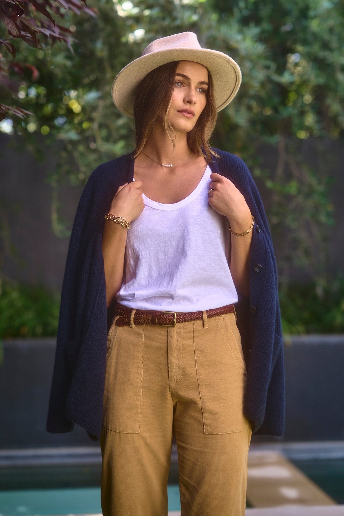   A woman stands outdoors wearing a white tank top, tan pants, and a hat, with the BRITT CARDIGAN by Velvet by Graham & Spencer draped over her shoulders. She gazes off into the distance. Trees and greenery are visible in the background, showcasing cozy and practical layering in cold weather. 